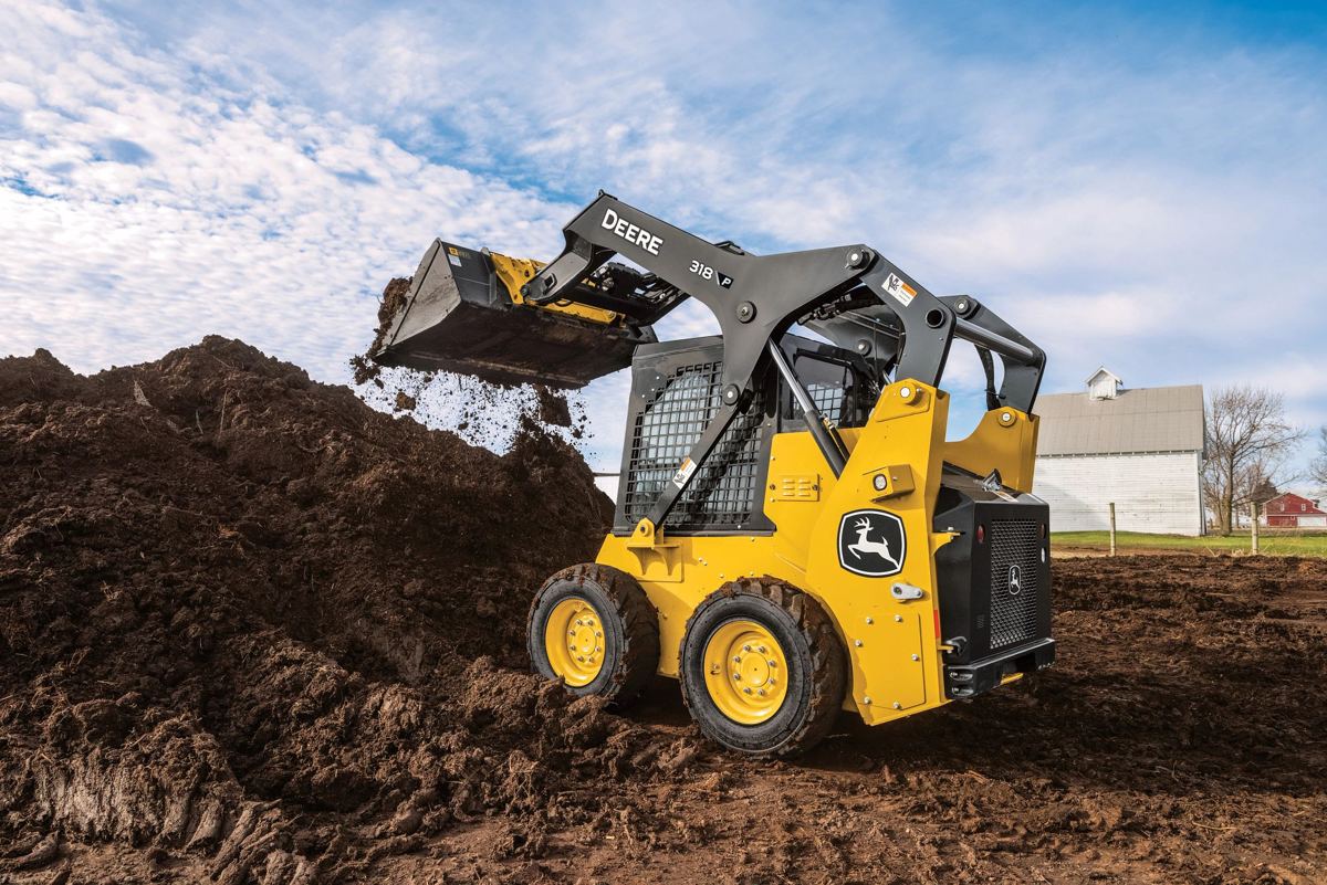 Yellow John Deere 318 P skid steer loader dumping dark brown dirt onto a large pile under a cloudy sky