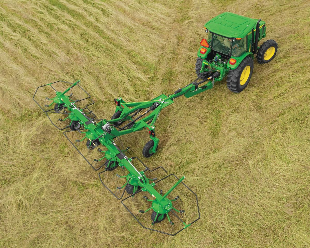 Green John Deere tractor pulling a large green hay rake through a field of cut hay