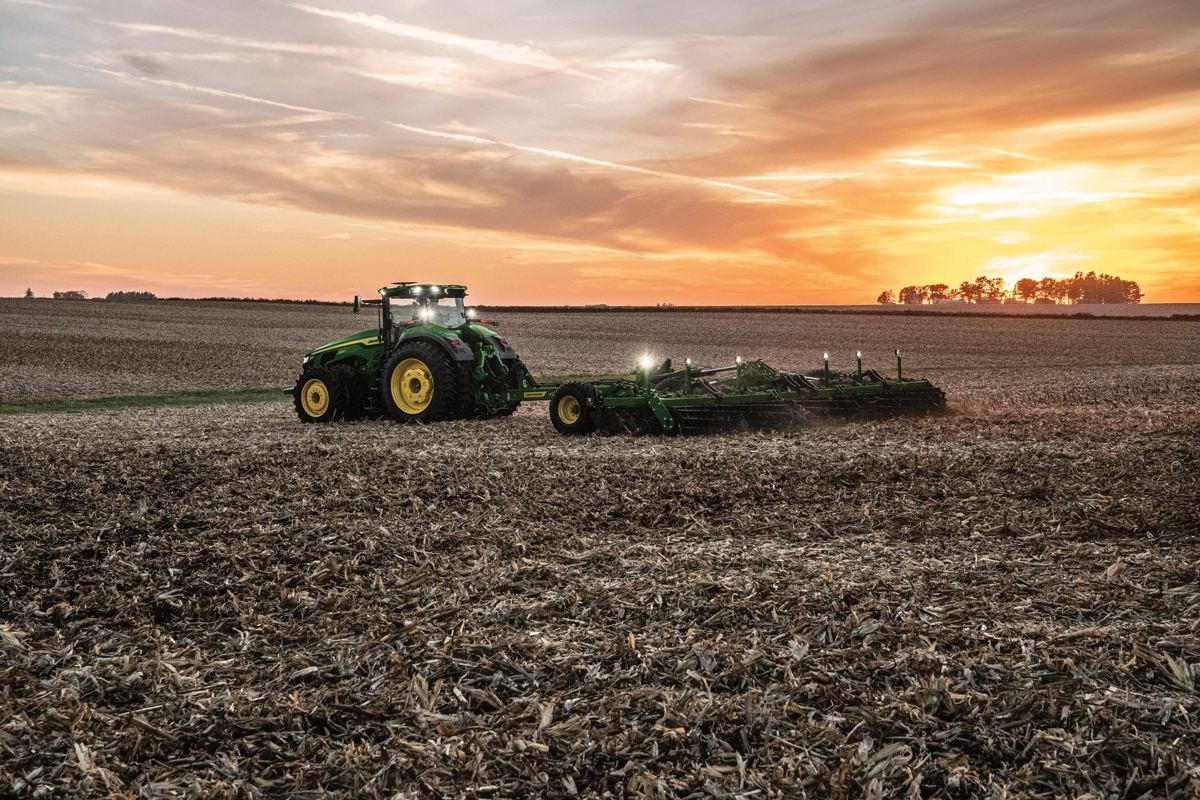 A green and yellow John Deere tractor with lights on pulls a tillage implement through a field of crop residue at sunset