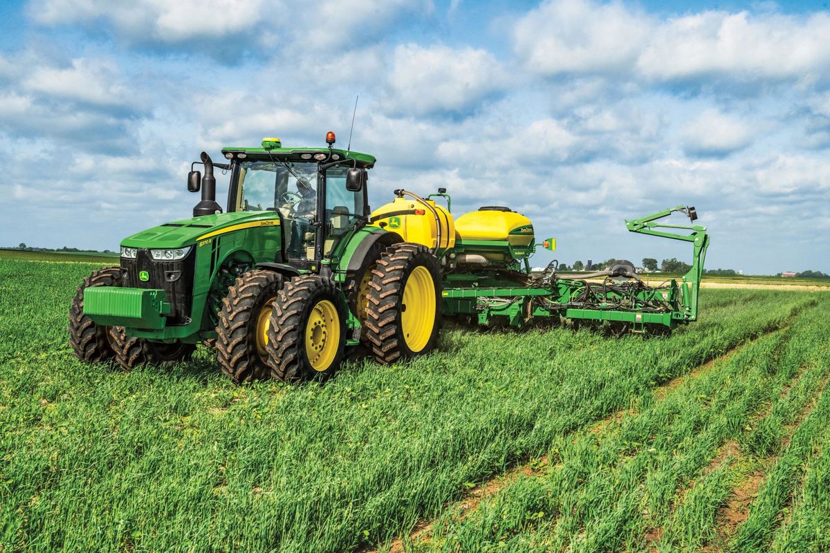A John Deere 8R series tractor with wheels is shown pulling a large planter, actively working in a green row crop field under a partly cloudy sky