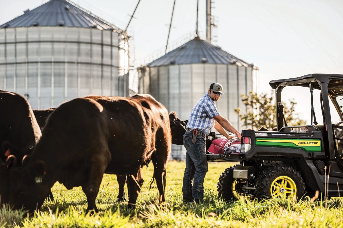 A man loading supplies into the cargo box of a John Deere full-size crossover Gator, with cows and grain silos in the background, depicting real farm work