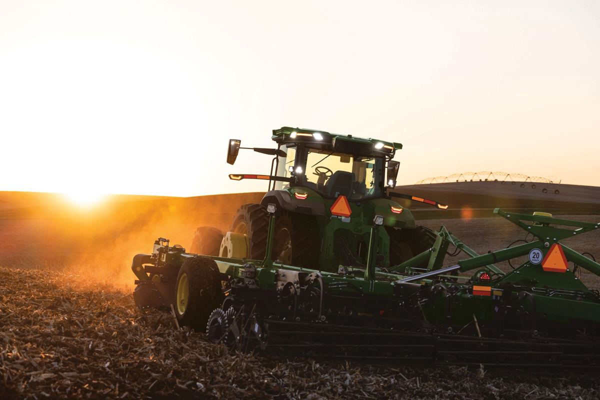A John Deere 2660VT vertical tillage tool is actively working corn or soybean residue behind a large John Deere tractor at sunset, with dust rising from the tilled soil
