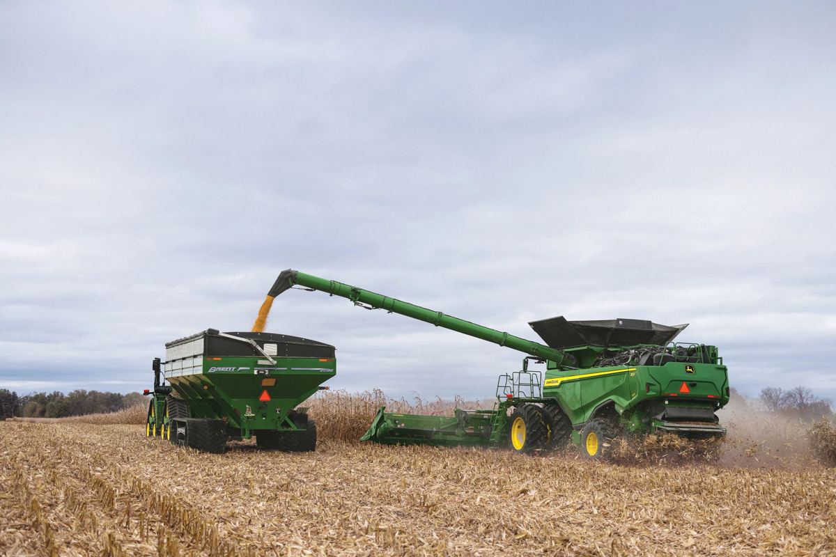 Green John Deere combine unloads golden grain via an auger into a green Brent grain cart in a stubble field