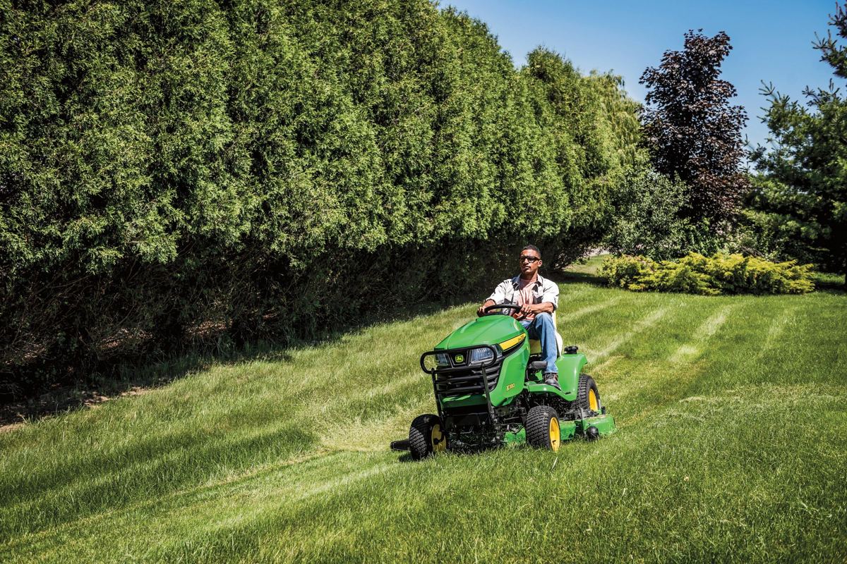 Man on a John Deere X380 riding lawn mower cutting grass in a large yard with trees