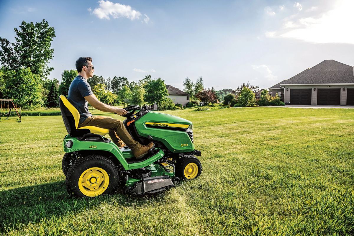 Man riding a green John Deere X384 lawn tractor on a large green lawn with houses and trees in the background