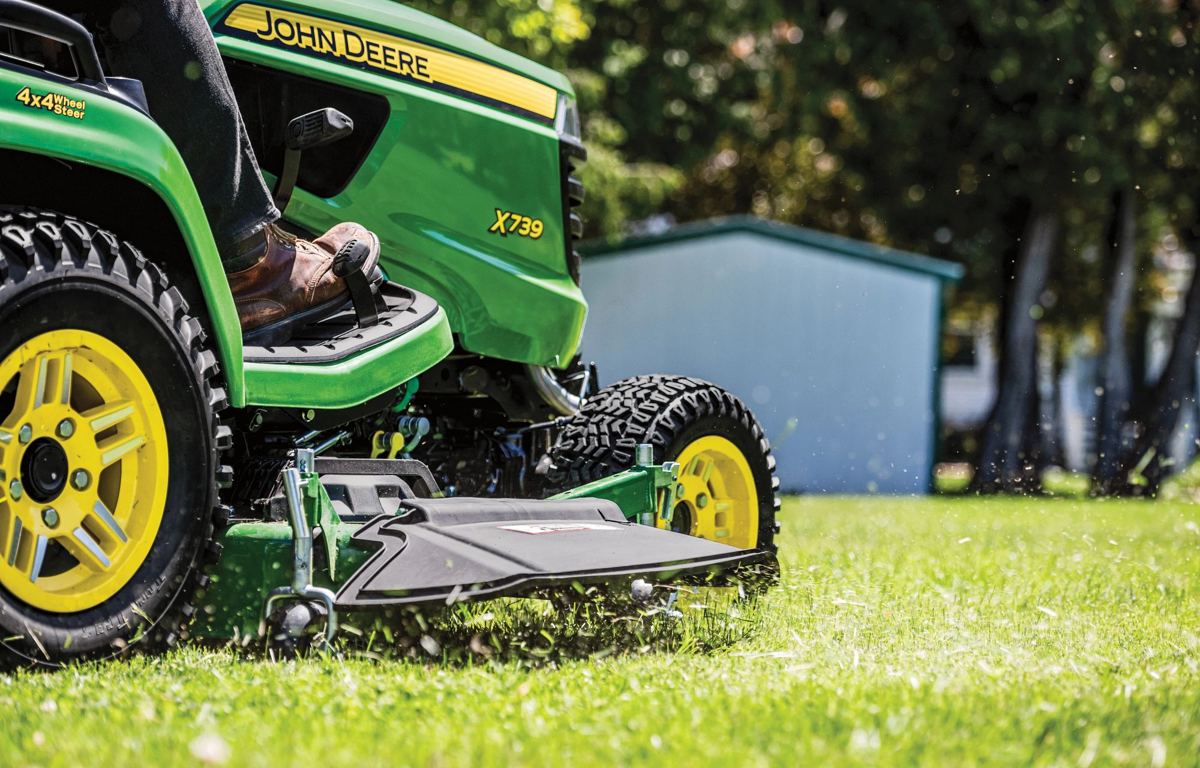 A John Deere X739 lawn tractor is actively mowing a green lawn, with grass clippings visible around the deck