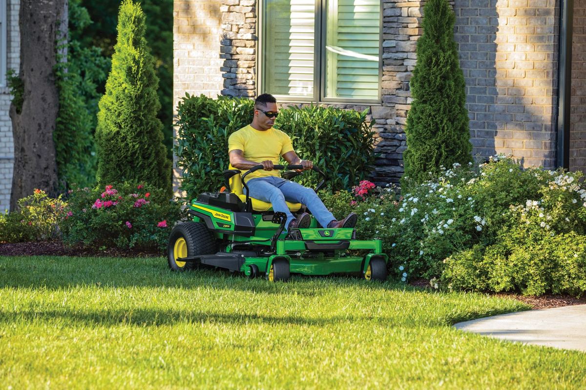 A person operates a John Deere Z300 ZTrak mower, cutting grass around a landscaped flower bed on a residential lawn