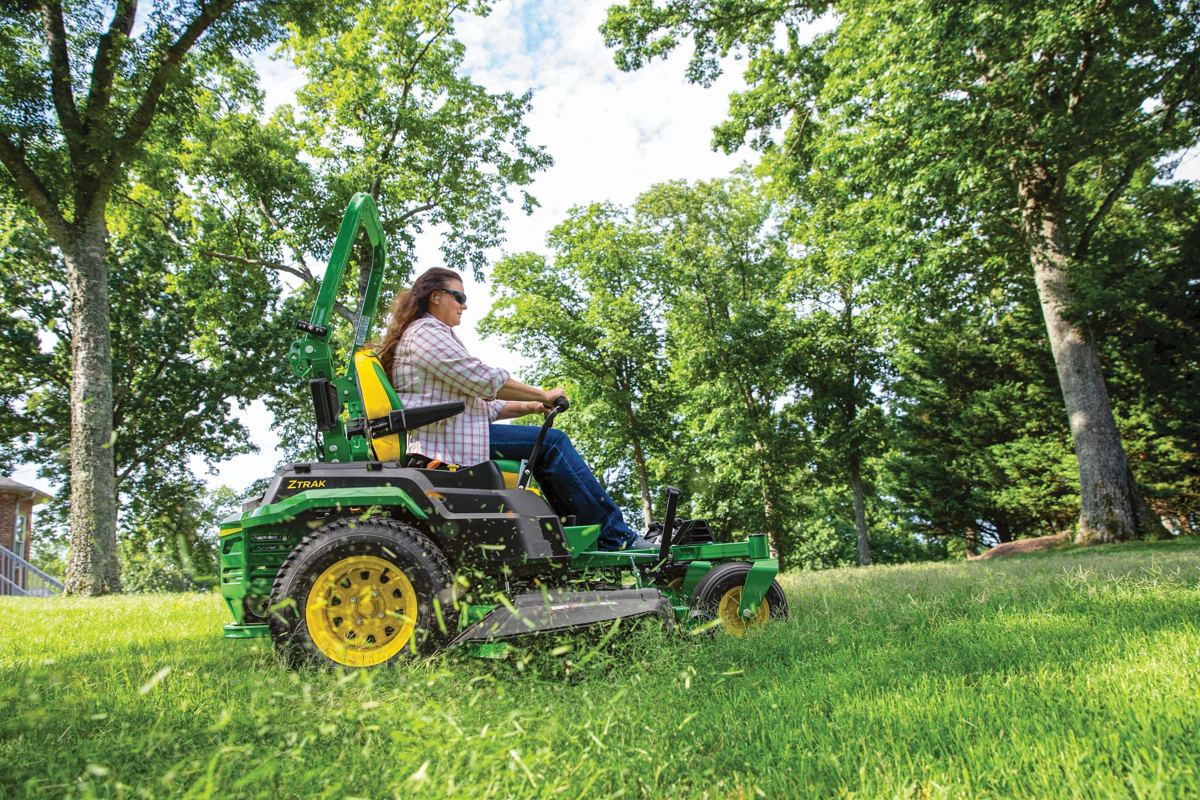 Woman in sunglasses driving a green John Deere ZTrak zero-turn mower, cutting grass in a sunny, treed yard