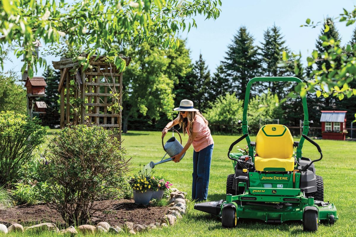 John Deere Z530M ZTrak zero-turn mower parked beside a flower bed while a homeowner waters plants