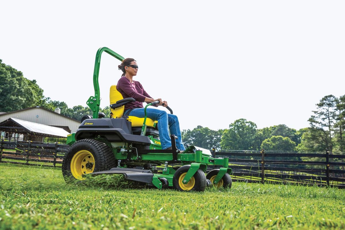 Woman on a John Deere ZTrak zero-turn mower cutting grass near a barn and fence