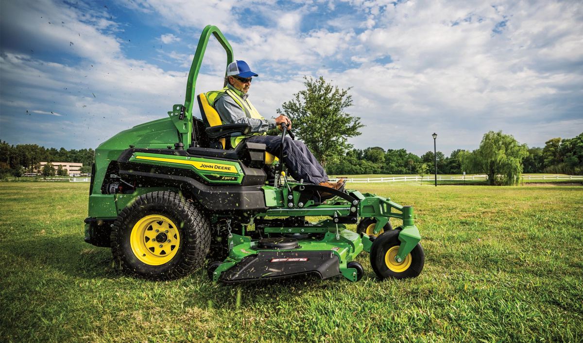 A person operates a John Deere ZTrak ride-on mower in a large, open grassy field under a blue sky, perfectly depicting the equipment mowing a vast open area