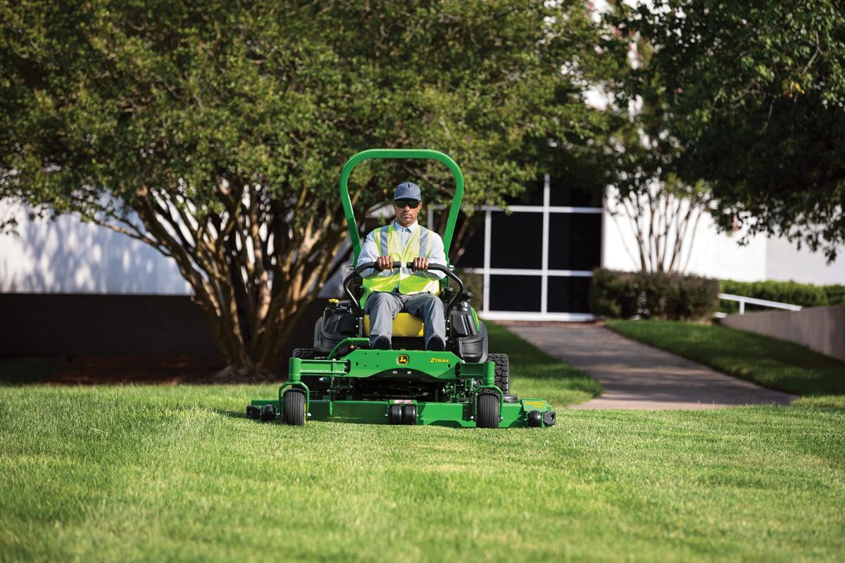 A landscaper operates a John Deere Z900 ZTrak zero-turn mower, actively mowing a well-maintained lawn in front of a commercial building
