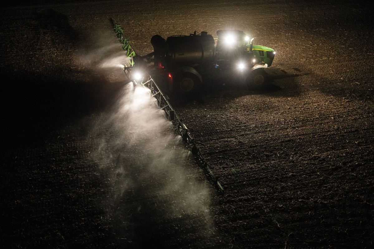 John Deere tractor with a wide sprayer boom applying liquid to a dark field at night, illuminated by its bright lights
