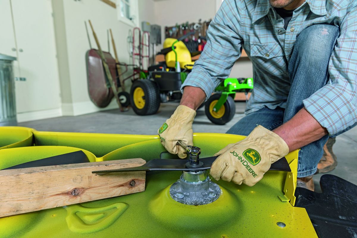 Man in John Deere gloves using a wrench to work on a lawnmower blade on a yellow deck in a garage