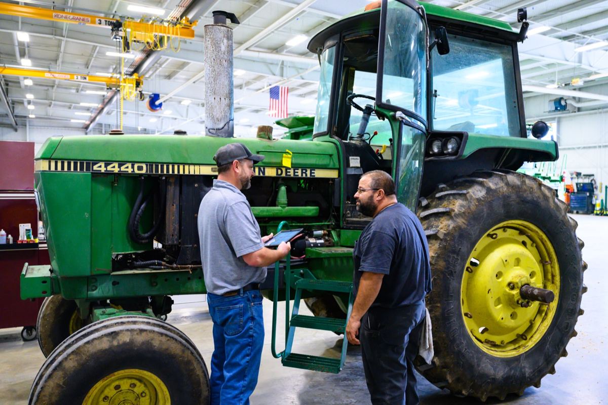 Two mechanics with a tablet discussing a John Deere 4440 tractor in a service bay