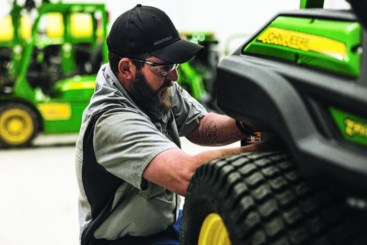 Man in safety glasses and Koenig cap working on a John Deere machine with other equipment in background