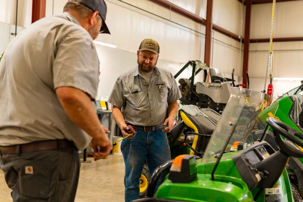 Two Koenig Equipment technicians with a measuring tape next to John Deere lawn mowers in a service bay