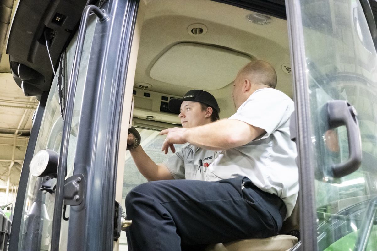 Two men in work shirts inside a large equipment cab, one pointing at an interior panel