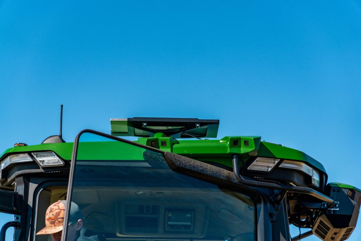 Close-up of a green John Deere tractor cab with a person inside, showing roof-mounted sensors and lights against a blue sky