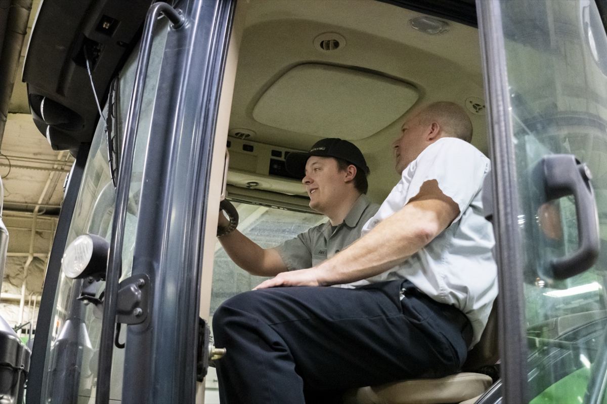 Two men in a large green equipment cab, one man pointing at an interior panel