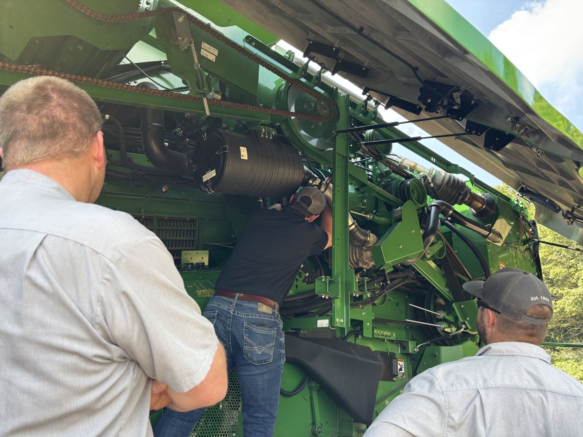 Three men inspecting and working on a large green agricultural machine with visible chains and hoses