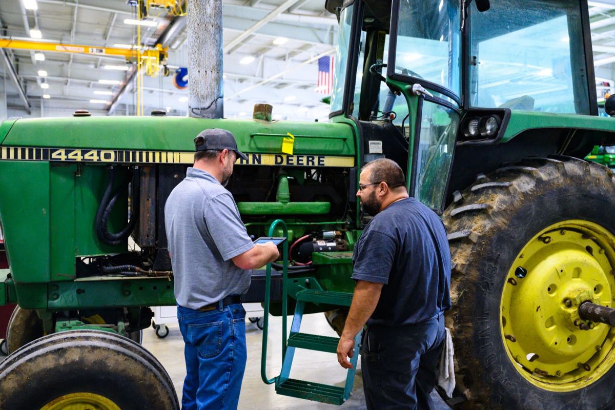 Two technicians inspecting a green John Deere 4440 tractor in a service bay