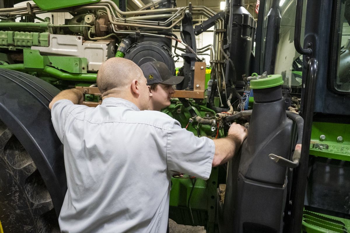 Two men, one in a John Deere cap, work on the engine of a green John Deere tractor