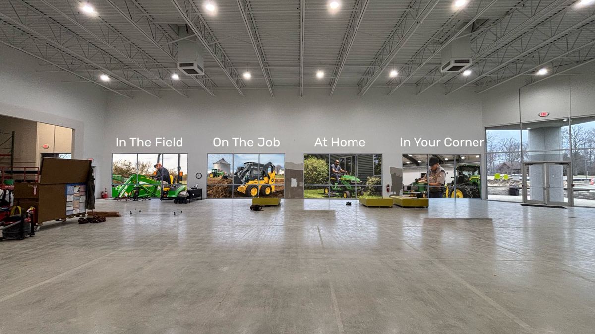 Empty showroom with four window displays featuring John Deere equipment for field, job, home, and service