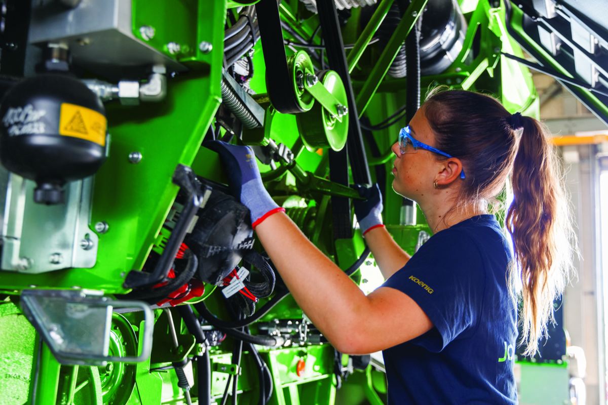 Koenig Equipment technician in safety glasses and gloves adjusting components and belts on a green John Deere machine