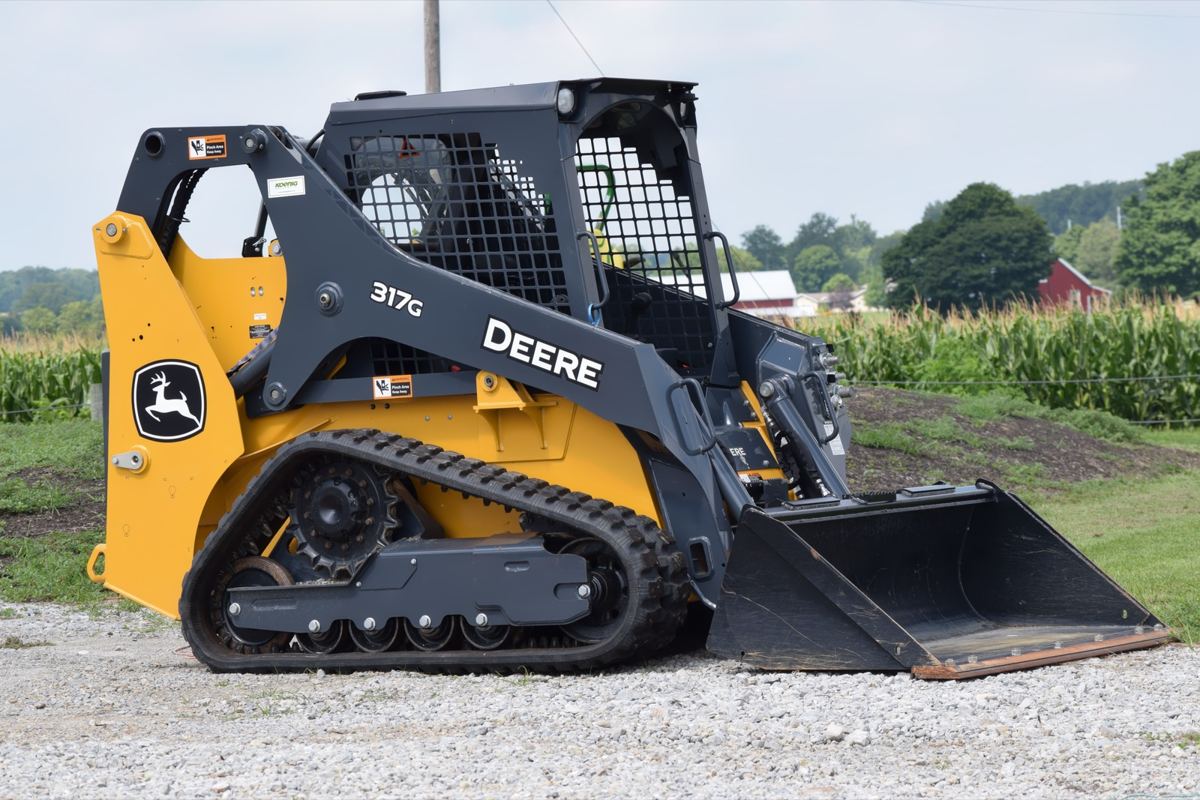 John Deere 317G compact track loader with a bucket attachment on a gravel lot beside a cornfield