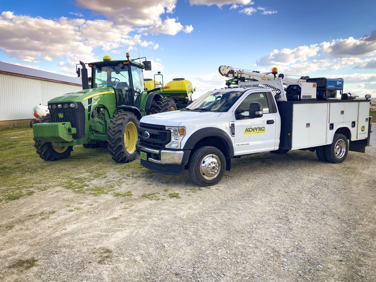 Green John Deere 8R tractor and white Koenig service truck with crane on a gravel lot