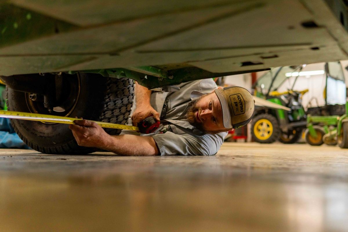 Koenig Equipment mechanic lying on the floor measuring under a green John Deere vehicle's tire