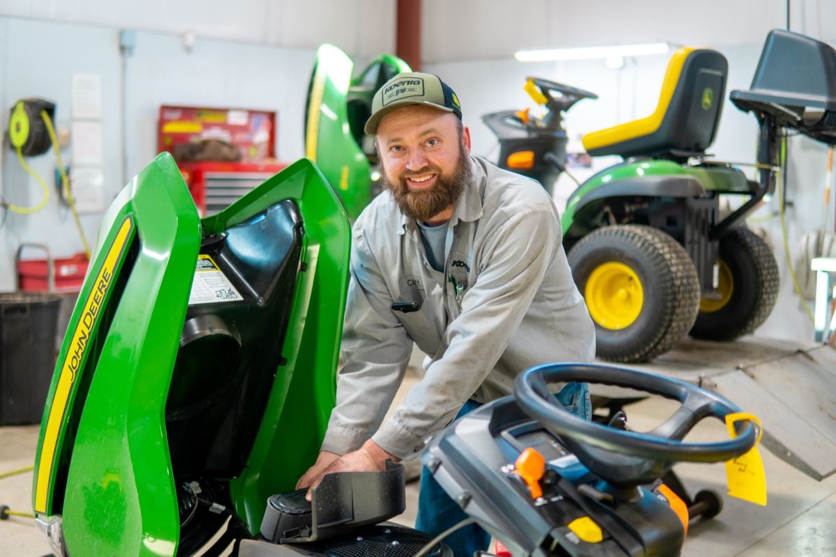 Smiling man in a work uniform and cap working on a green John Deere riding mower with its hood open in a service bay