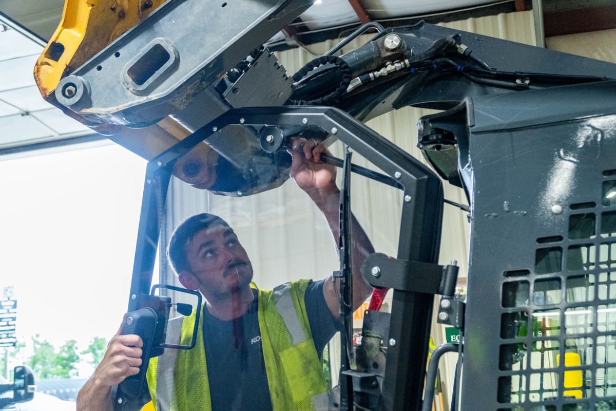 Technician in safety vest working on the overhead arm of a John Deere skid steer loader