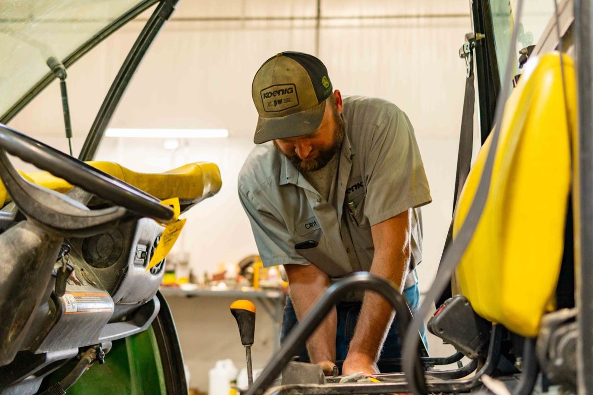 Mechanic Clint in a Koenig hat and shirt inspecting a John Deere utility vehicle's engine
