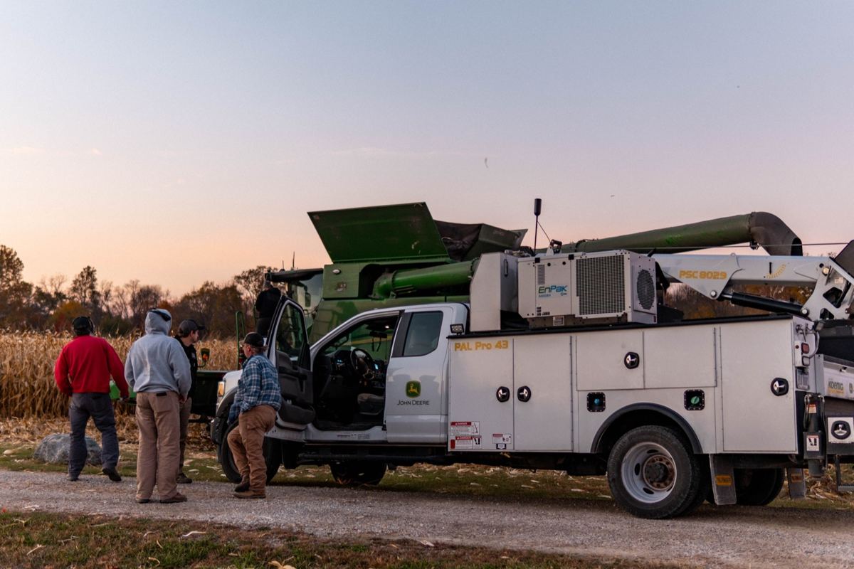 Four men stand by a John Deere service truck with a PAL Pro 43 crane and a green combine harvester in a cornfield