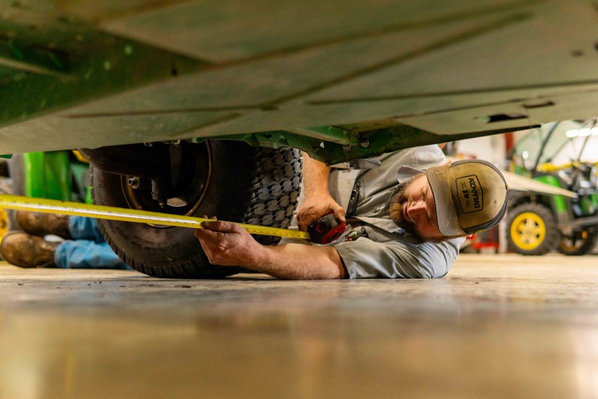 A man in a Koenig hat lies on his back under a green vehicle, measuring with a yellow tape