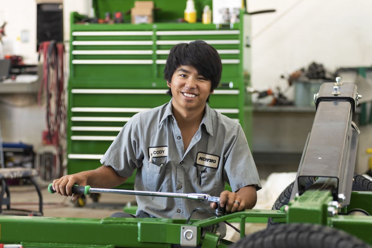 Smiling man in a Koenig work shirt using a wrench on green John Deere equipment in a service bay