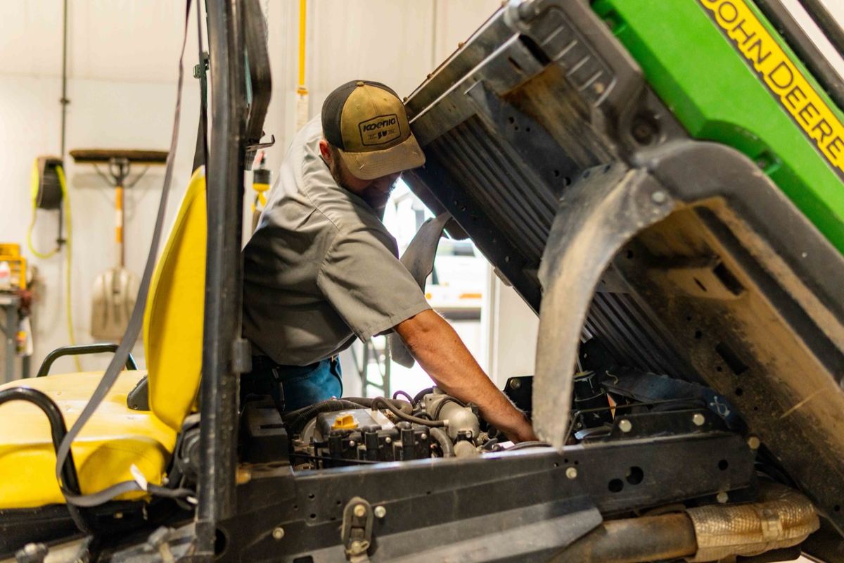 Man in Koenig hat servicing the engine of a green and yellow John Deere utility vehicle