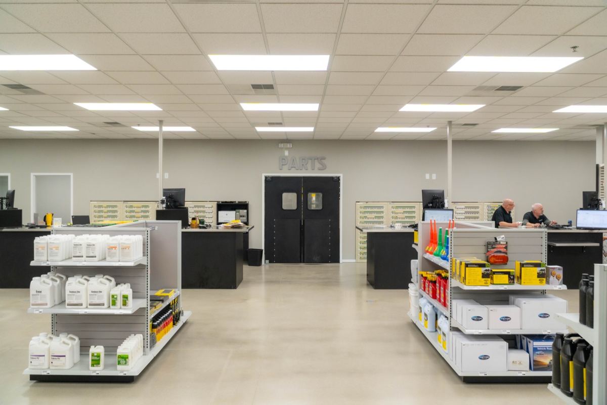 Interior of a parts department with product shelves, service counters, two men working, and a 'PARTS' sign above double doors
