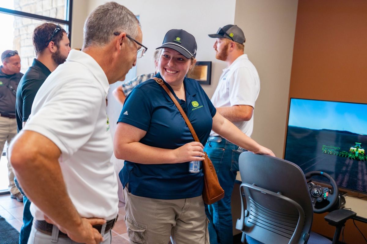 A man talks to a smiling woman in a John Deere shirt and cap next to a farming simulator