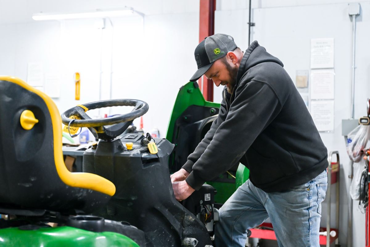 Man in John Deere cap working on the engine of a green and black John Deere riding mower