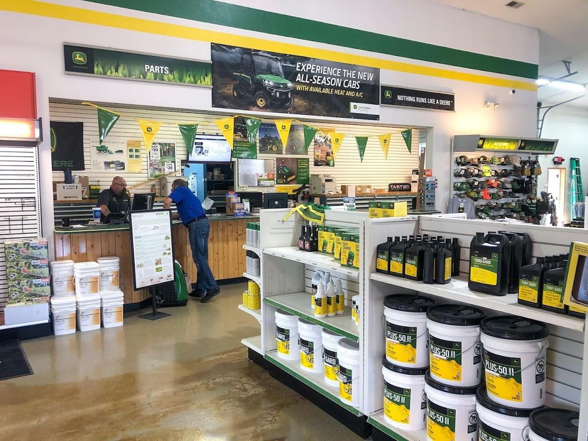 Two men at a John Deere parts counter, surrounded by shelves of John Deere oils, lubricants, and banners