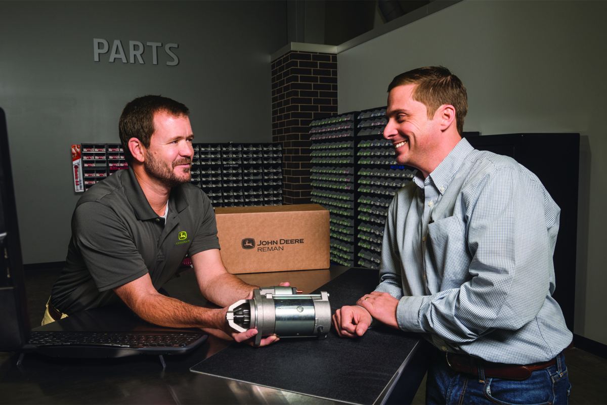 An employee is holding a John Deere REMAN starter motor at a parts counter, representing a part being handled at a dealership parts counter