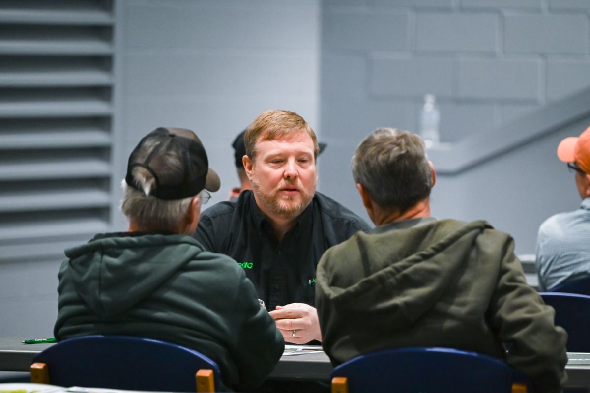 Bearded man in black shirt talking to two men in hoodies at a table, backs to camera