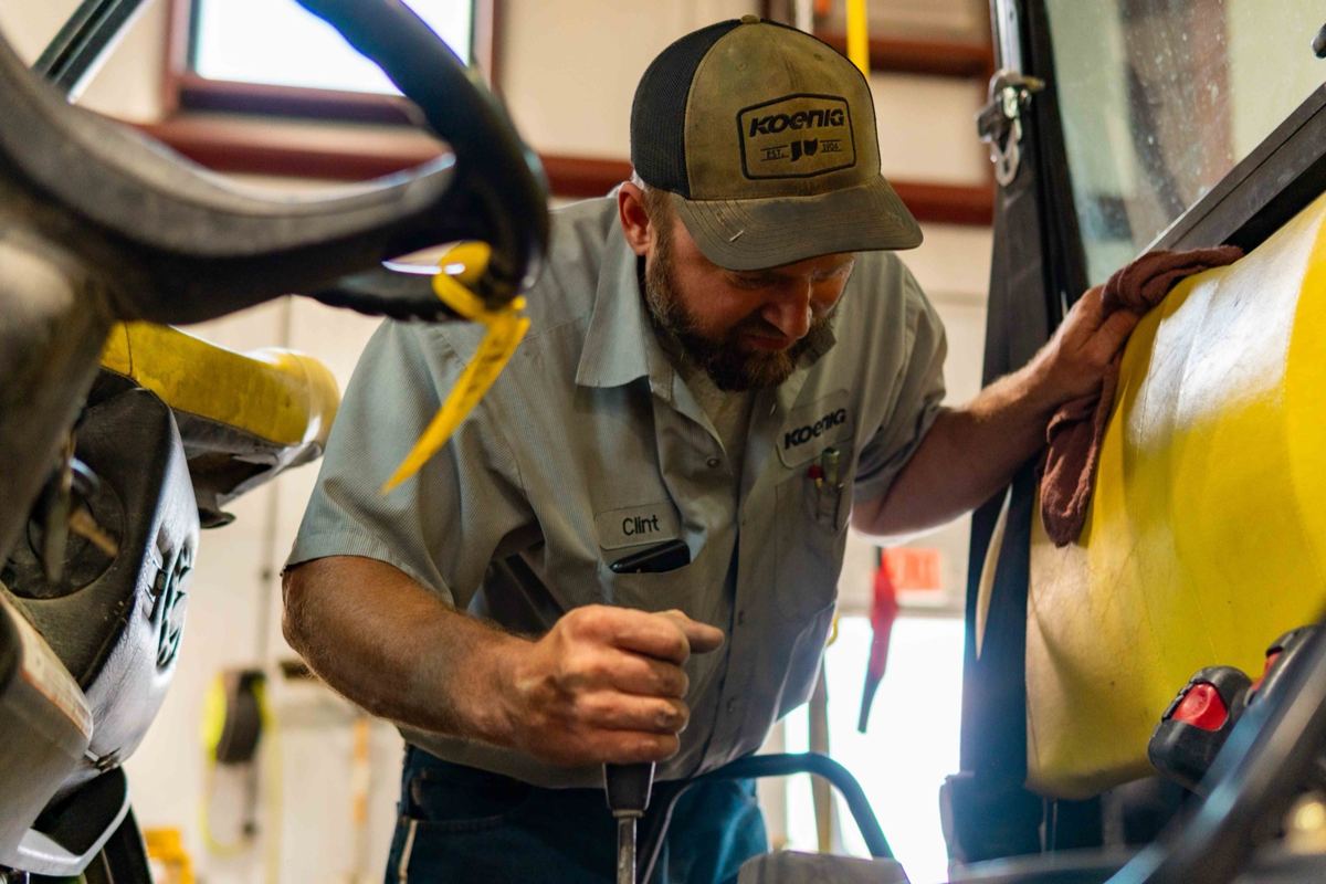 Koenig technician Clint working on yellow machinery, holding a tool and wiping a surface with a rag