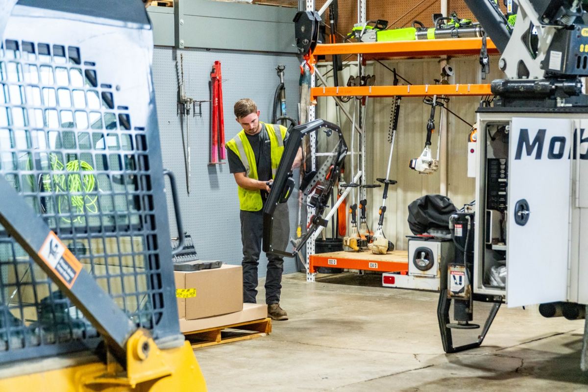 Technician in a safety vest assembling equipment in a workshop with power tools, a yellow skid steer, and a utility truck