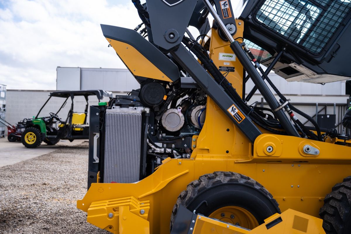 Close-up of a yellow skid steer loader with its engine compartment open, and a John Deere Gator in the background
