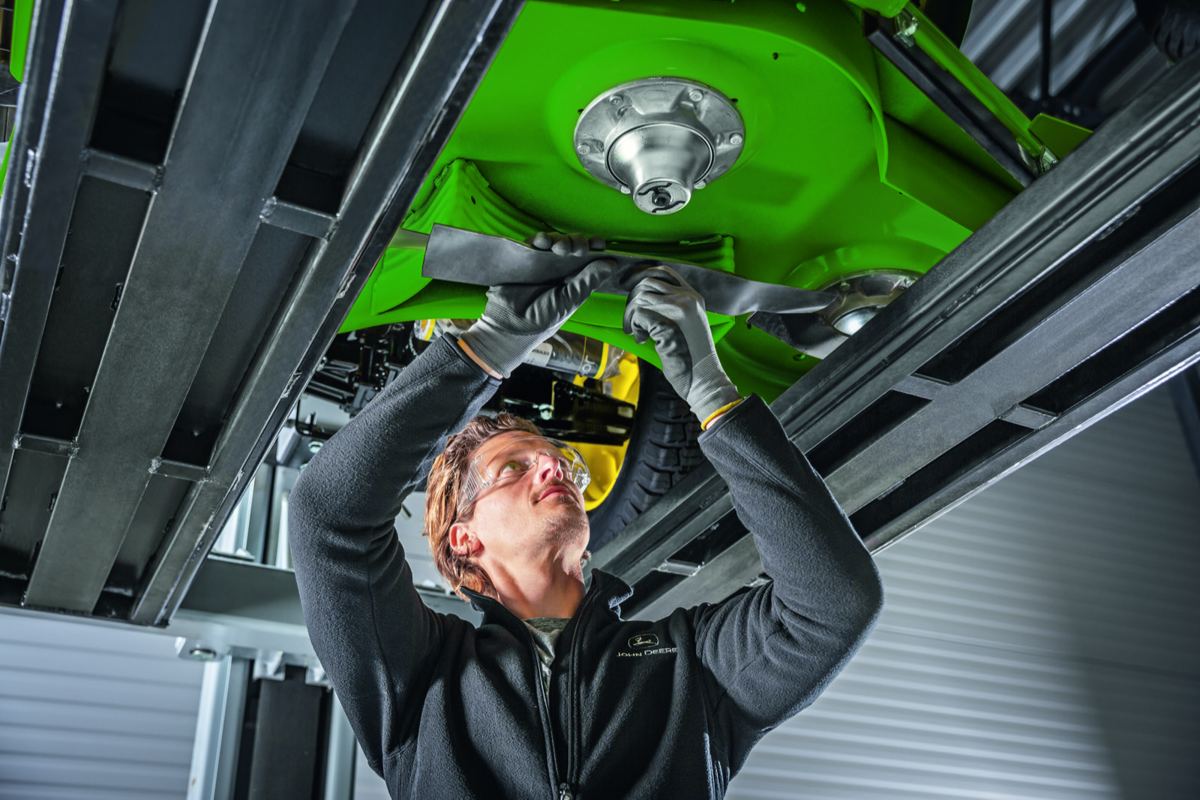 Technician in safety glasses and John Deere jacket working on a green mower deck on a lift