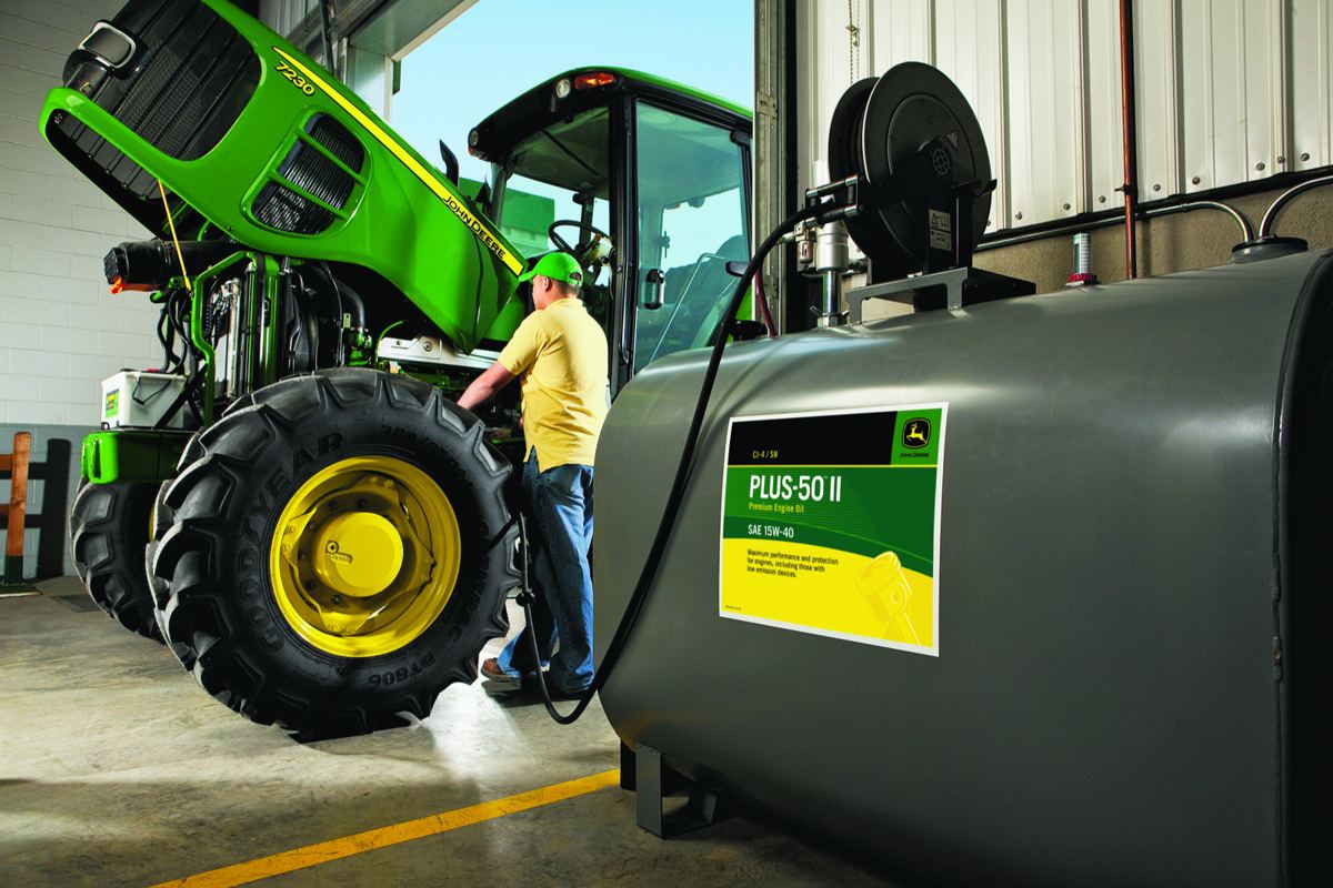 Man servicing a John Deere 7230 tractor with its hood open, next to a tank of John Deere PLUS-50 II engine oil
